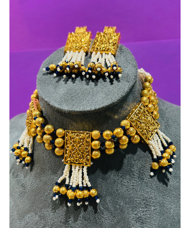 An ornate gold-toned jewelry set displayed on a gray mannequin bust against a purple background. The set includes a beaded necklace with rectangular filigree-style gold pendants, matching drop earrings, and white and navy blue bead tassels.