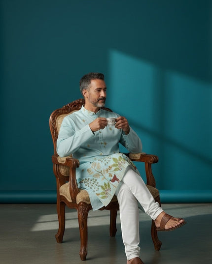 Kurti Connection USA light blue silk kurta with intricate embroidery and white pajama, worn by a man sitting in a chair.